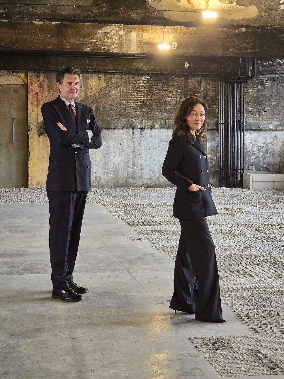 Stefano Rabolli Passera and Marisa Chearavanont stand confidently in a raw industrial space, both dressed in elegant dark attire, with overhead lighting casting a warm glow on the concrete floor and exposed brick wall behind them.