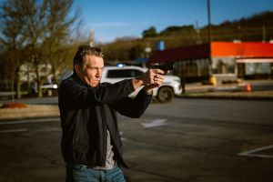 A man in civilian clothes holds a handgun in a two-handed grip, aiming intently in a parking lot, with a police vehicle and fast food restaurant in the background.
